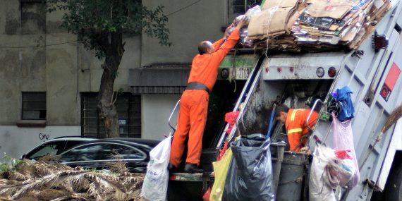 man in orange jacket and blue denim jeans standing near garbage