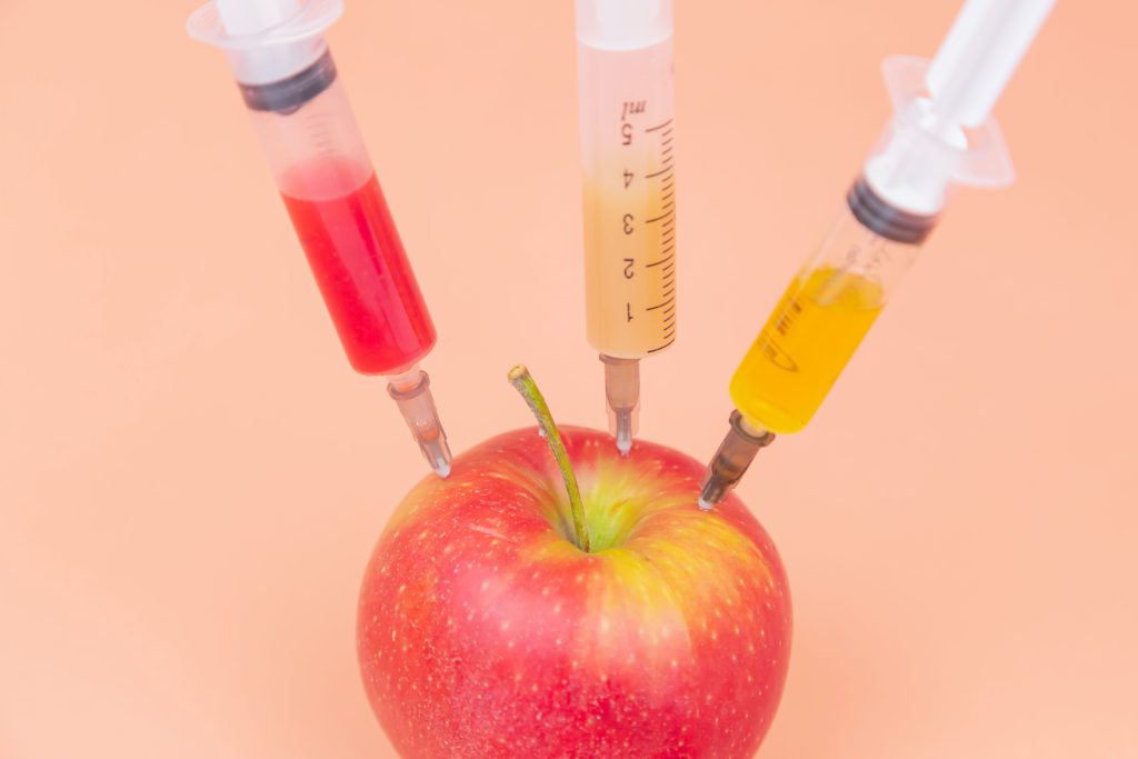 Syringes with different chemicals injections into a red apple on an orange background. GMO. Chemistry. Poison. Fruits. Place for an inscription. The close plan. Drugs.