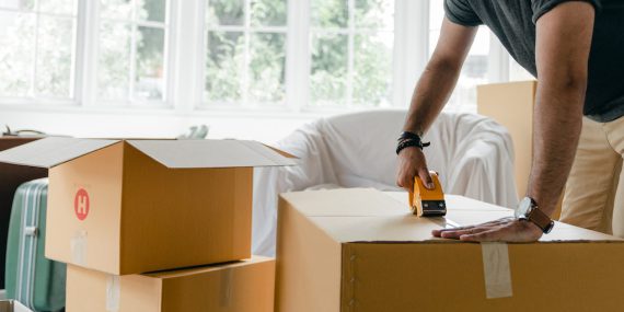 Crop unrecognizable man in casual clothes packing carton box using scotch tape dispenser for moving personal items to new apartment
