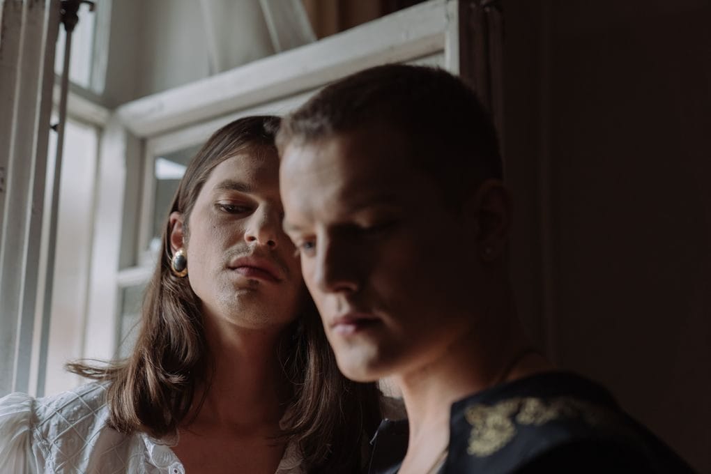 Dramatic indoor portrait of two androgynous individuals by a window, evoking mystery and sophistication.