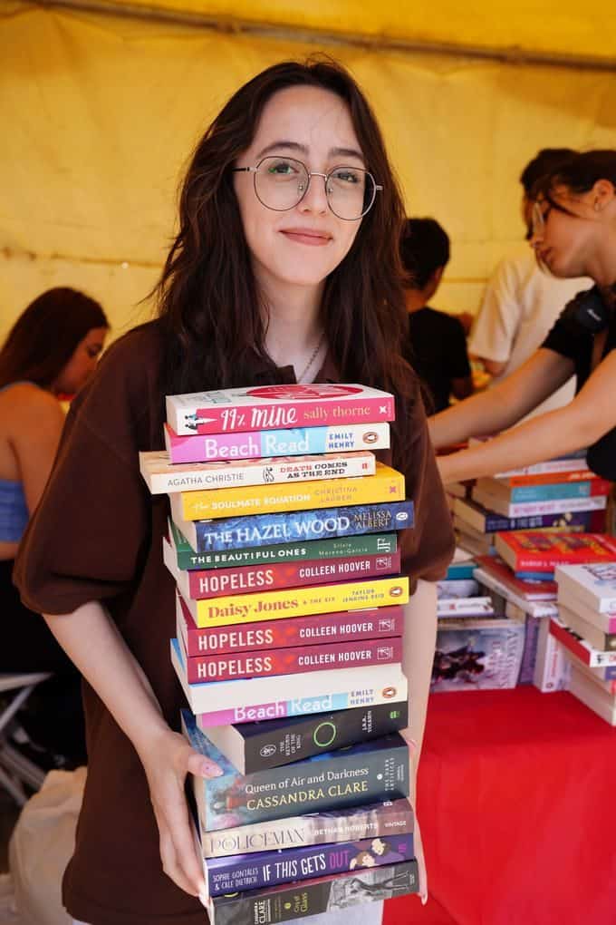 A young woman in glasses smiles while holding a large stack of popular books at an indoor event.