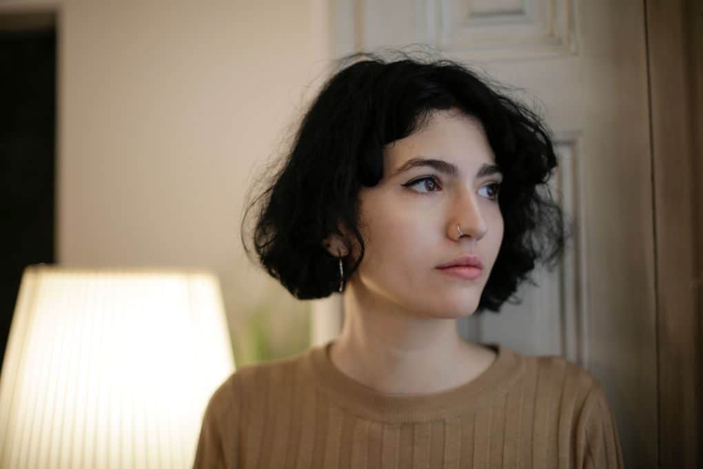 A thoughtful woman with short, wavy hair and a nose ring gazes away in a softly lit room.